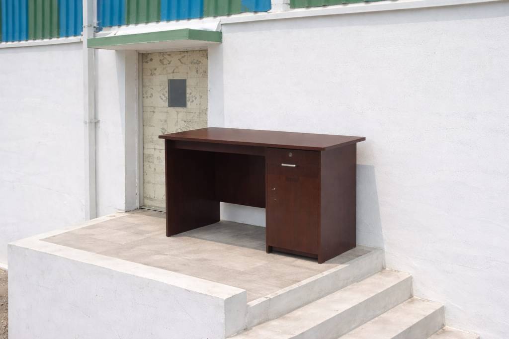 A wooden desk placed on a concrete platform next to a door with a small window, surrounded by a white wall and colored metal roofing.